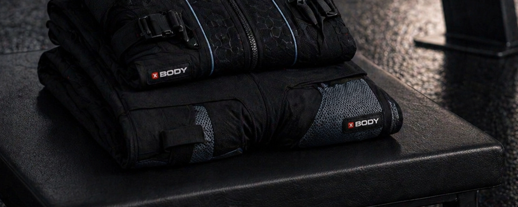 XBody Zwei zusammengefaltete schwarze Trainingswesten mit XBODY-Logos auf einer schwarzen Bank.
