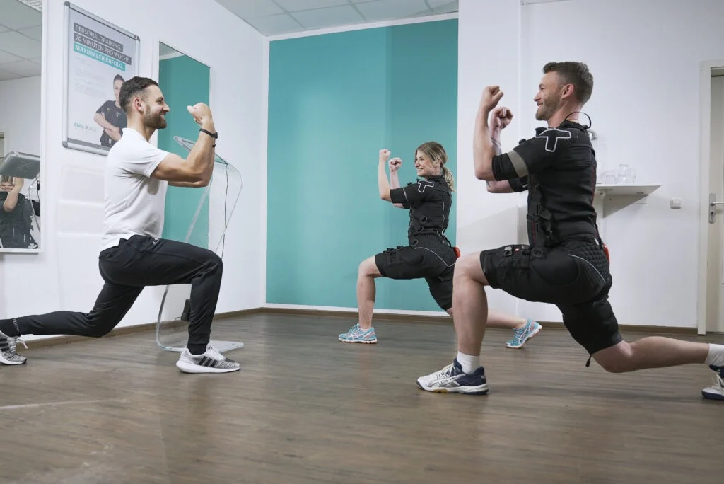 XBody Zwei Personen in EMS-Anzügen machen Ausfallschritte, während ein Trainer in einem Fitnessstudio eine Übung vorführt. XBody Zwei Personen in EMS-Anzügen machen Ausfallschritte, während ein Trainer in einem Fitnessstudio eine Übung vorführt.