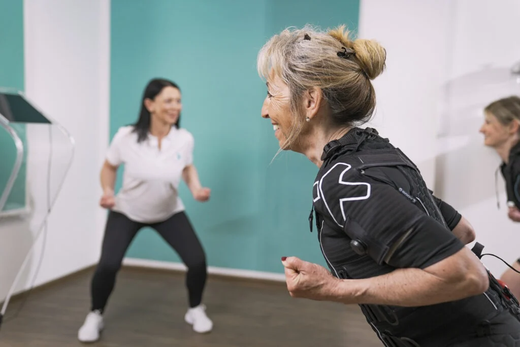 XBody Zwei Frauen, die drinnen Sport treiben; eine trägt Sportkleidung mit Sensoren, beide lächeln. XBody Zwei Frauen, die drinnen Sport treiben; eine trägt Sportkleidung mit Sensoren, beide lächeln.