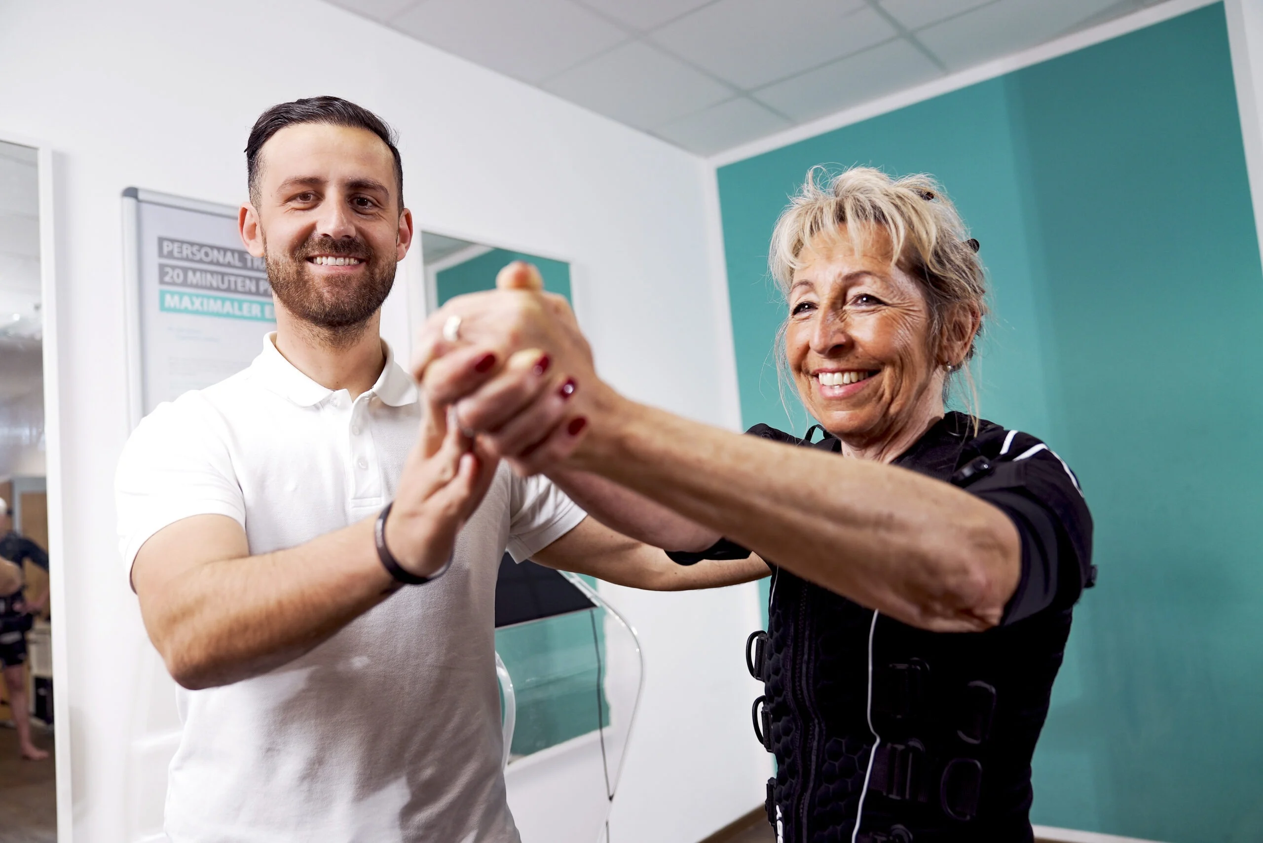 XBody Eine lächelnde ältere Frau und ein Mann, die in einer Physiotherapie- oder Fitnessumgebung Händchen halten. XBody Eine lächelnde ältere Frau und ein Mann, die in einer Physiotherapie- oder Fitnessumgebung Händchen halten.
