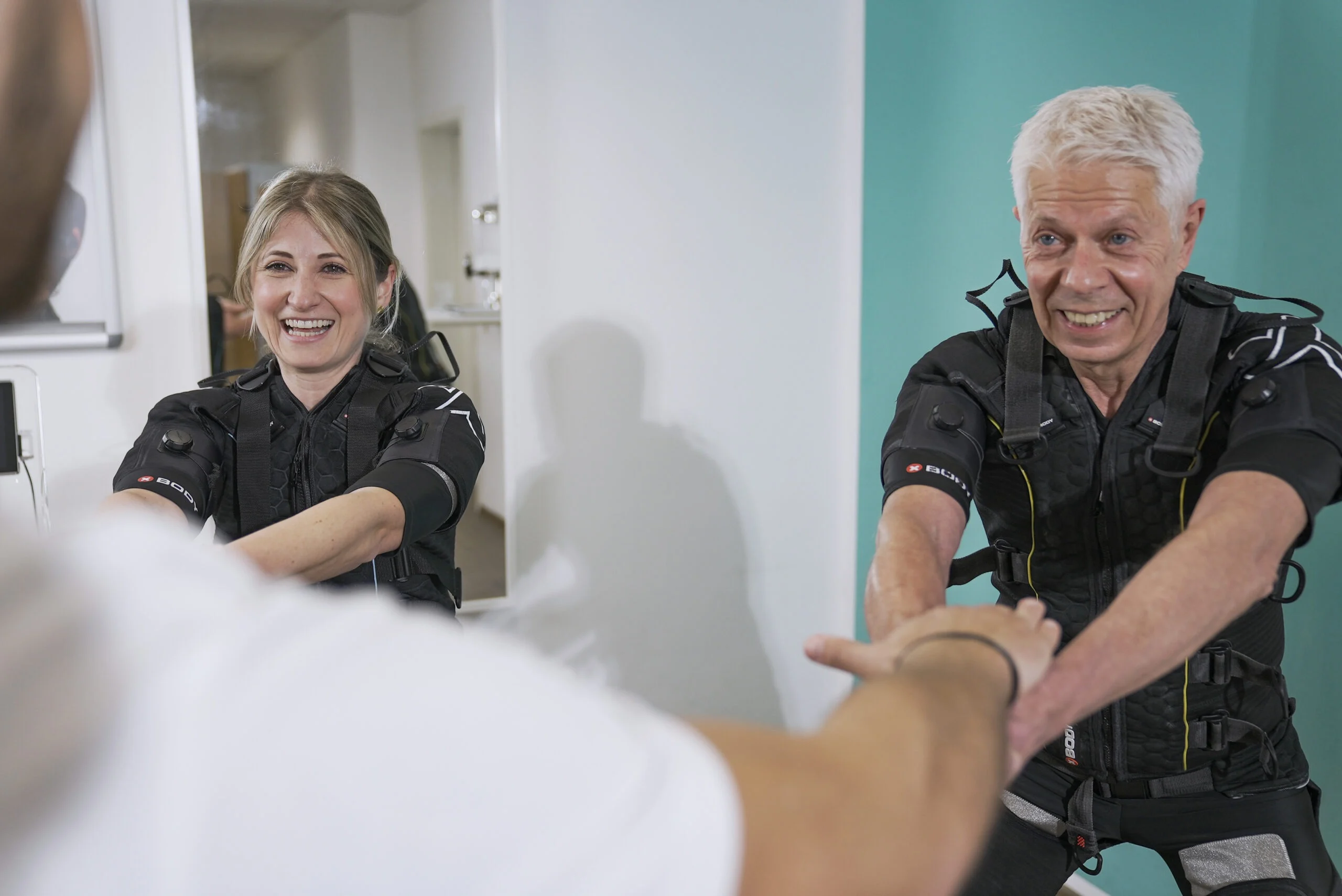 XBody Zwei Personen in Sportkleidung, die lächeln und sich gemeinsam mit einem Trainer in einem hellen Raum dehnen. XBody Zwei Personen in Sportkleidung, die lächeln und sich gemeinsam mit einem Trainer in einem hellen Raum dehnen.