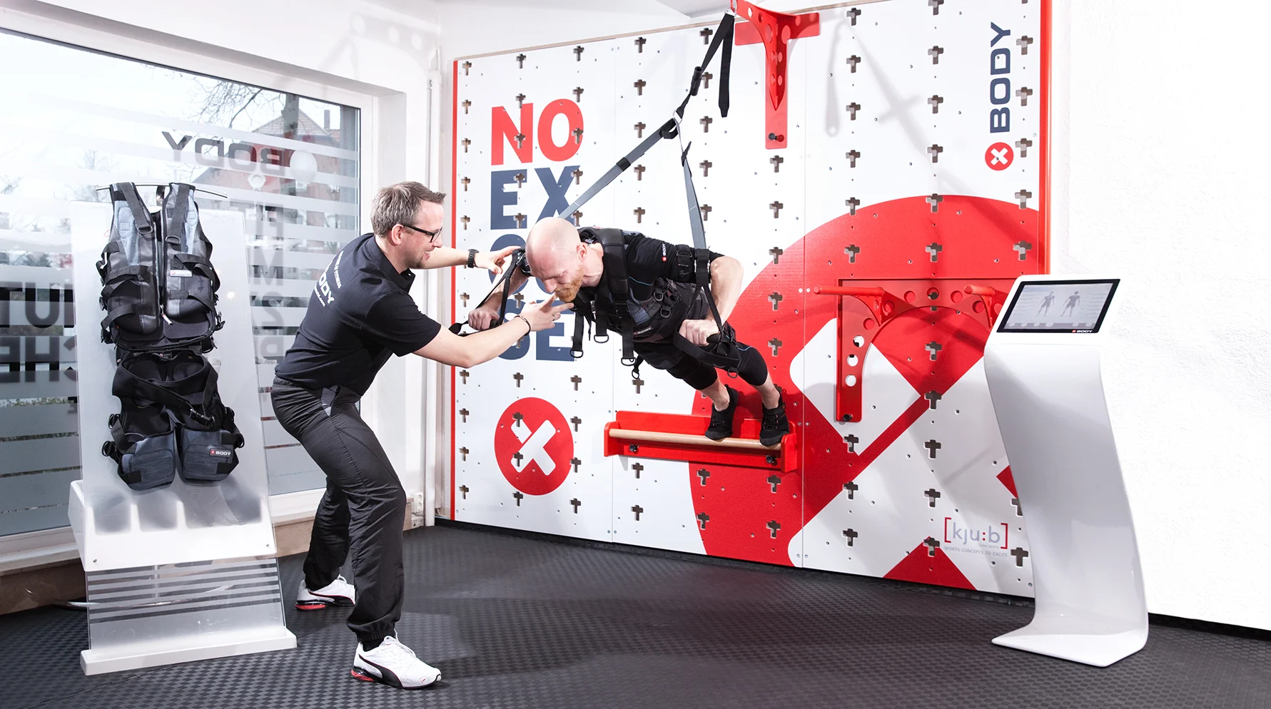 Bild Gründung XBody Training Germany 2015 XBody Ein Trainer unterstützt einen Mann, der in einem Gurtzeug an einem hängenden Trainingssystem in einem Fitnessstudio hängt.