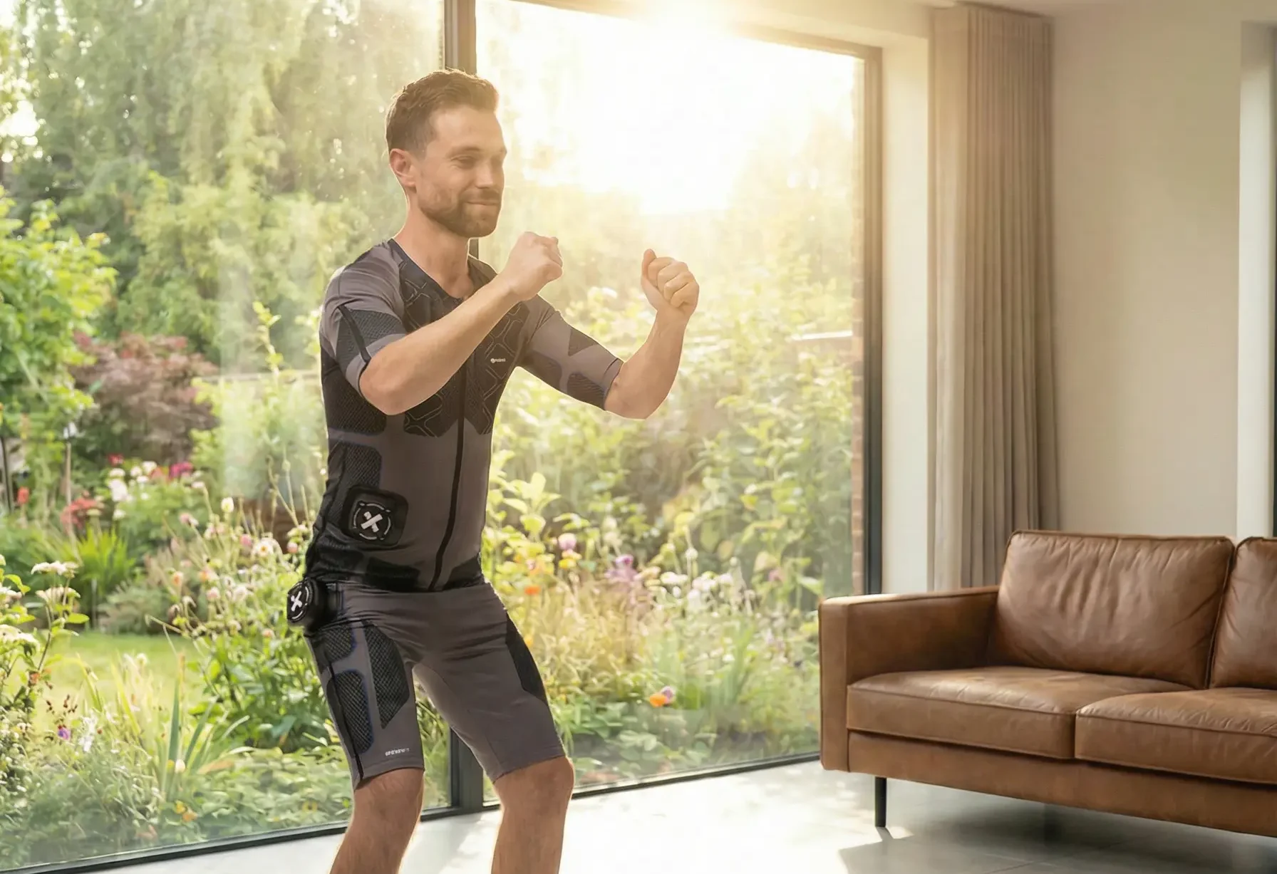 Training im Wohnzimmer copy XBody Ein Mann in Sportkleidung trainiert drinnen vor großen Fenstern mit Blick auf den Garten und in der Sonne.